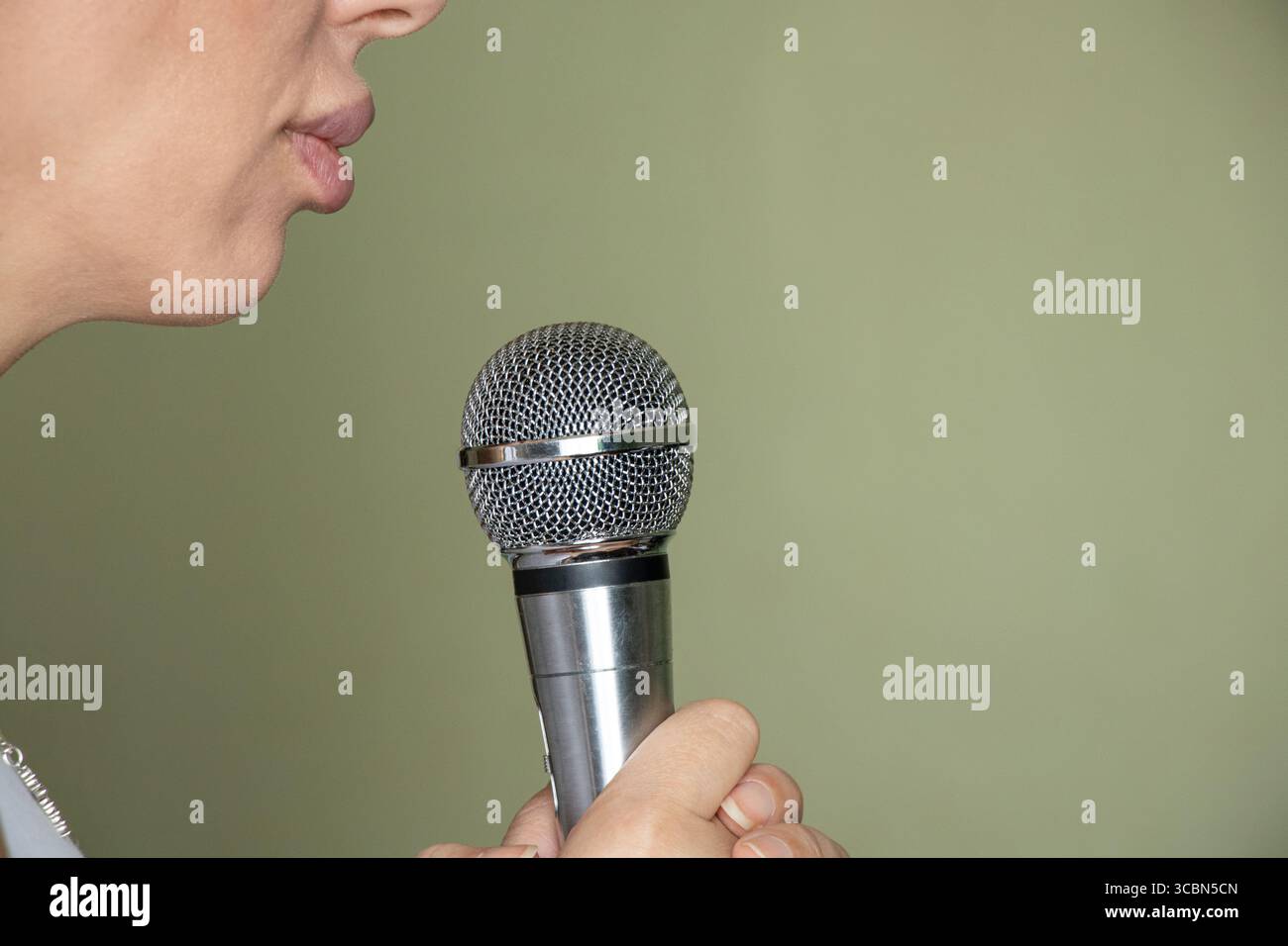 Woman holding microphone in front of mouth, close up of face and hand, preparing to sing or perform on green background at home Stock Photo