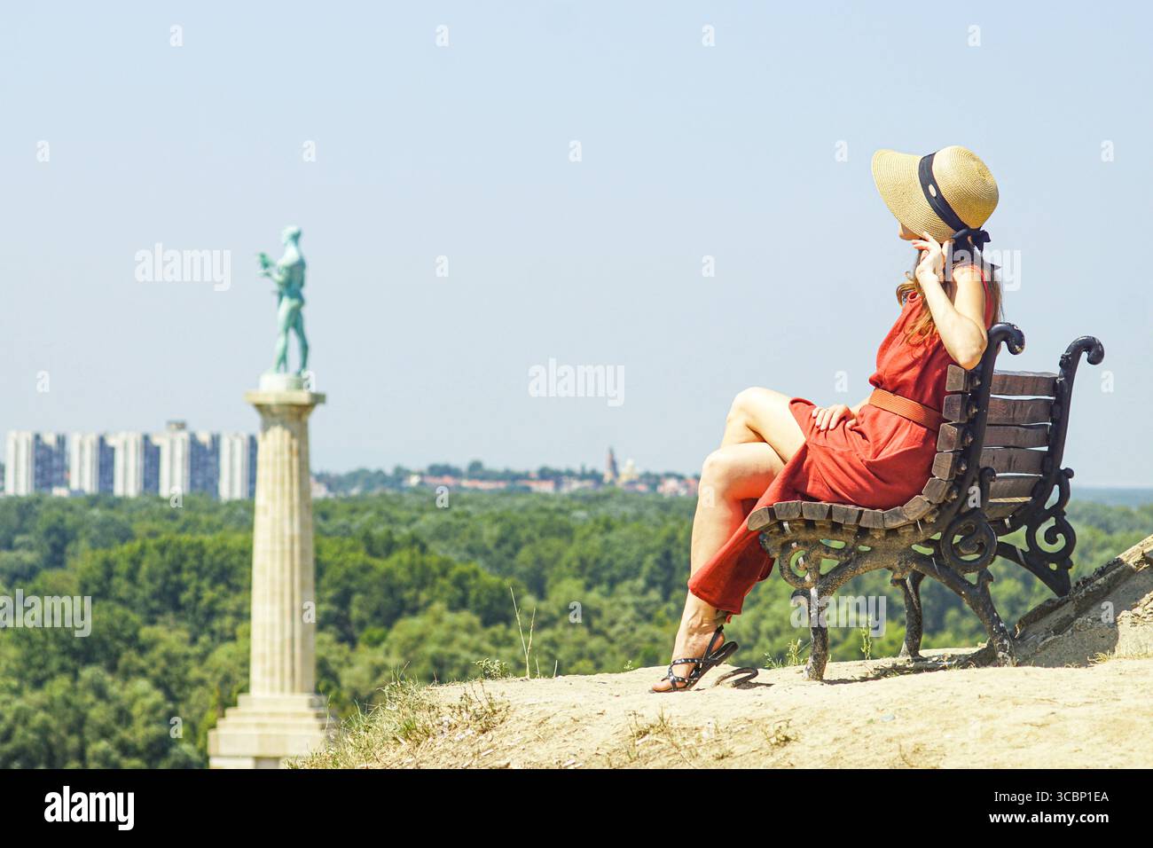 Slow travel: a woman in a red dress and hat sits on a bench at Kalemegdan fortress, gazing towards the Pobednik monument and New Belgrade skyline Stock Photo