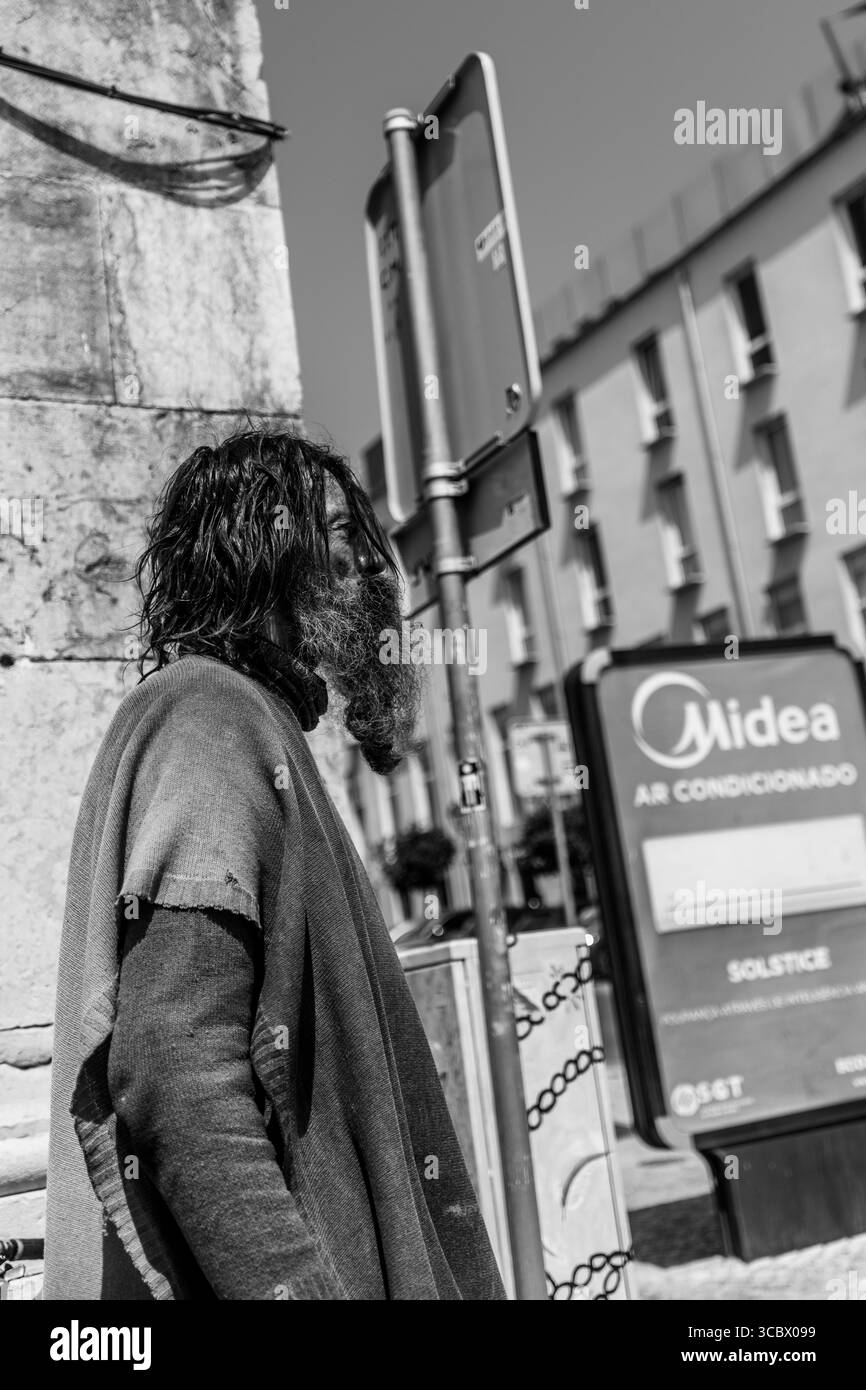 Algarve, Portugal – 2025.06.28 A man with a long beard and unkempt hair stands in the sun wearing a tattered poncho, blending into the urban backdrop Stock Photo