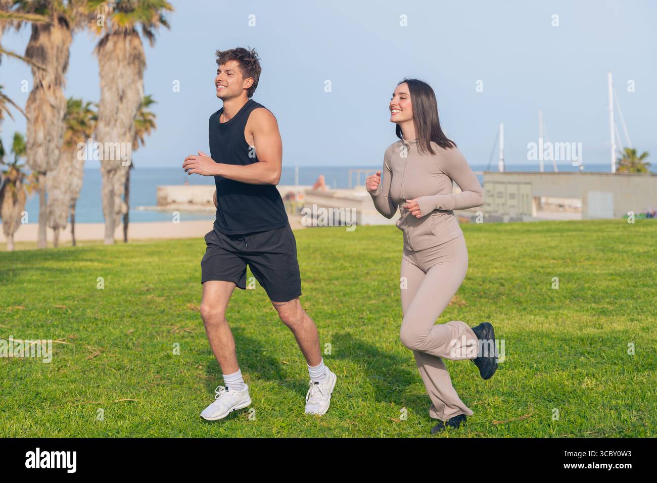 Fit couple jogging together on seaside lawn, smiling and chatting Stock Photo