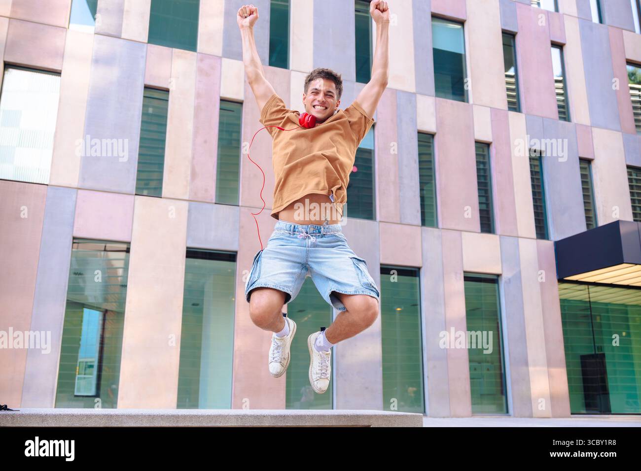 Happy young man jumping outdoors with arms raised in front of modern building Stock Photo