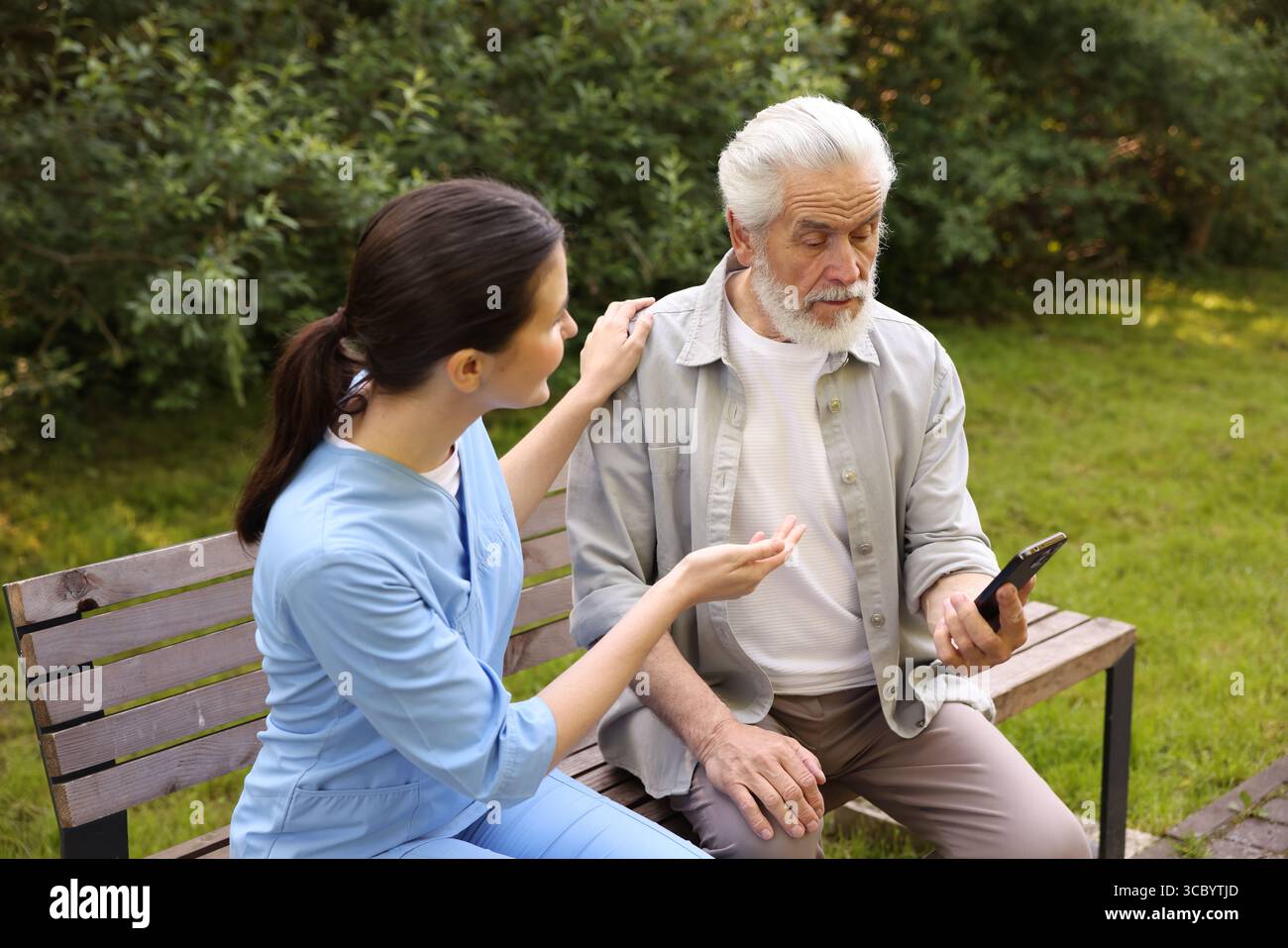 Nurse and elderly man using smartphone on bench outdoors Stock Photo