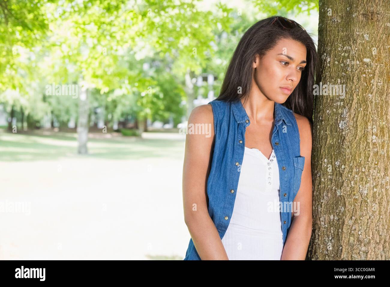 Woman leaning against tree trunk in sunlit park, gazing toward right with sunglasses, copy space Stock Photo