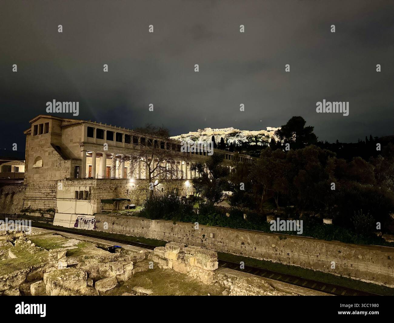 Roman Basilica ruins with graffiti-covered subway passing by, Ancient Agora and Acropolis in the background, Athens, Greece at night. - Smartphone Captured Stock Image