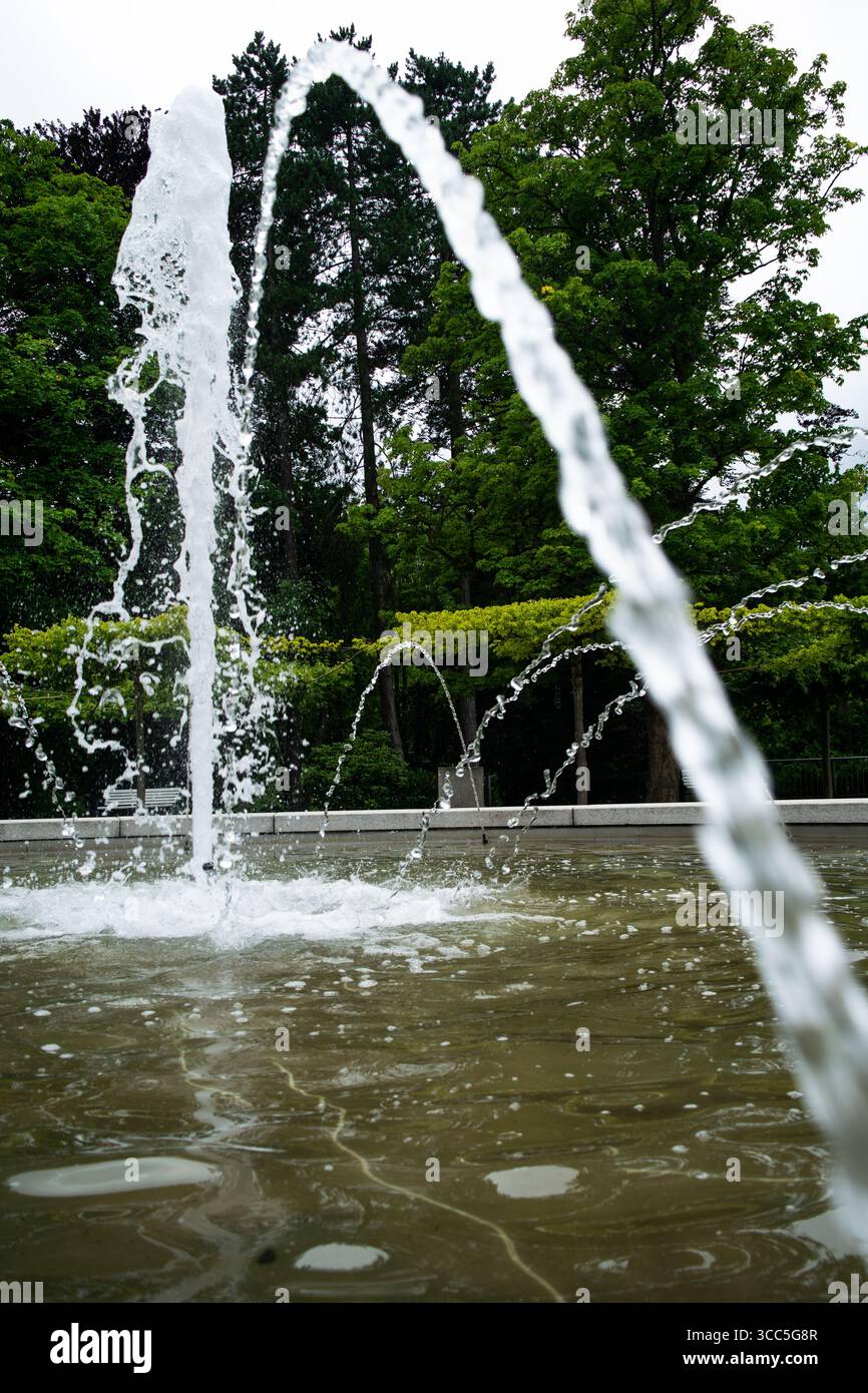 A refreshing stream of water arcs gracefully from a fountain in a peaceful public park, surrounded by vibrant green foliage. Stock Photo
