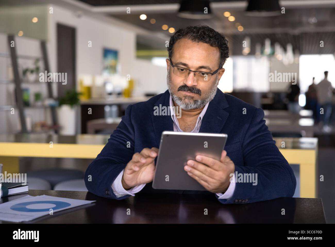Serious elder Indian executive man typing on tablet at table Stock Photo