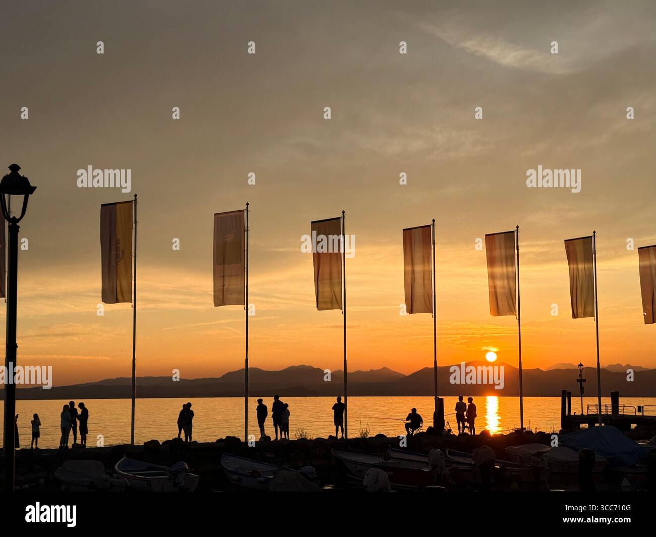 Cisano (near Bardolino) on Lake Garda, Italy. People gathered on harbour by flags for the sunset. - Smartphone Captured Stock Image
