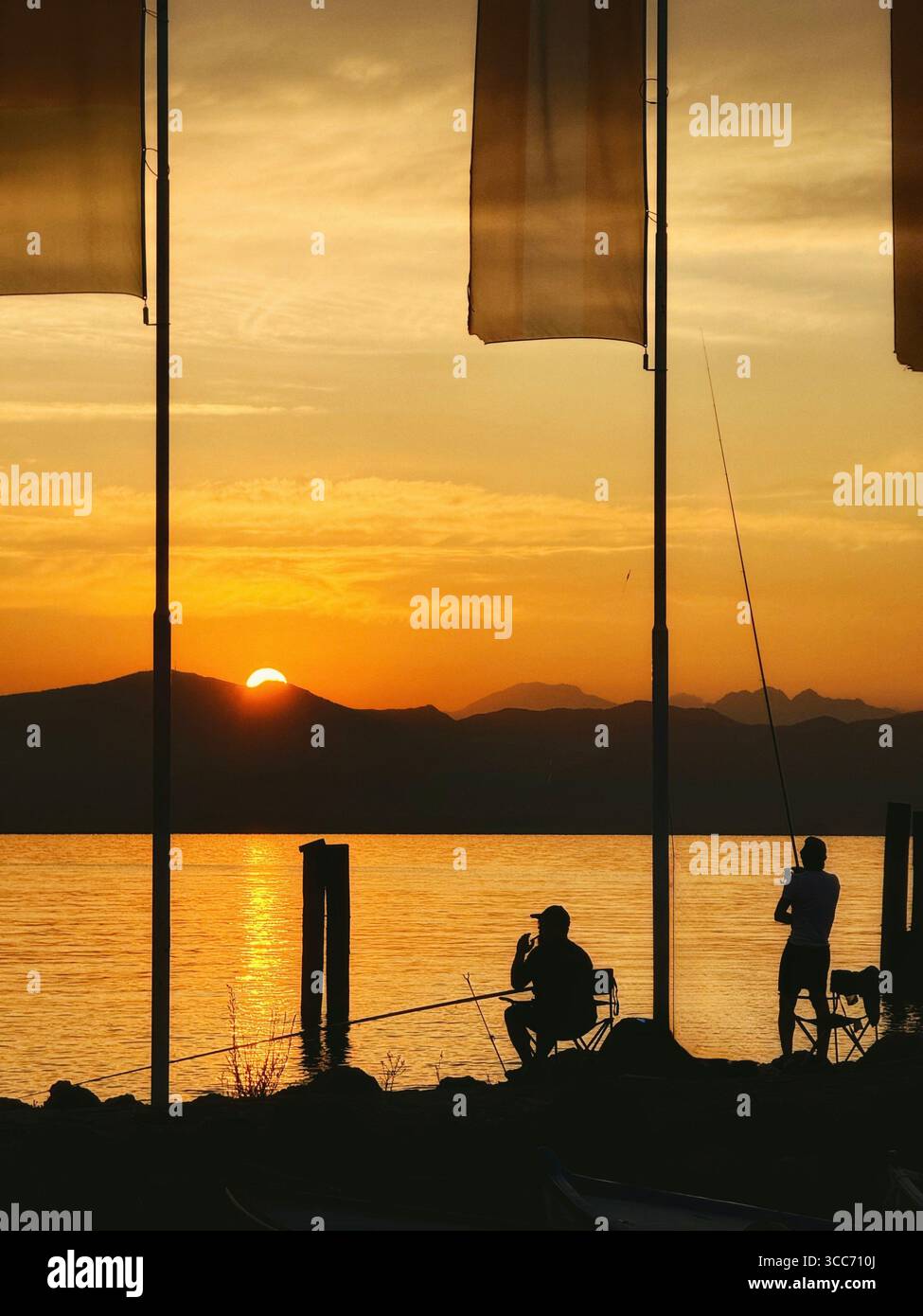 Cisano (near Bardolino) on Lake Garda, Italy. Two Fishermen in silhouette on harbour by flags at sunset. - Smartphone Captured Stock Image