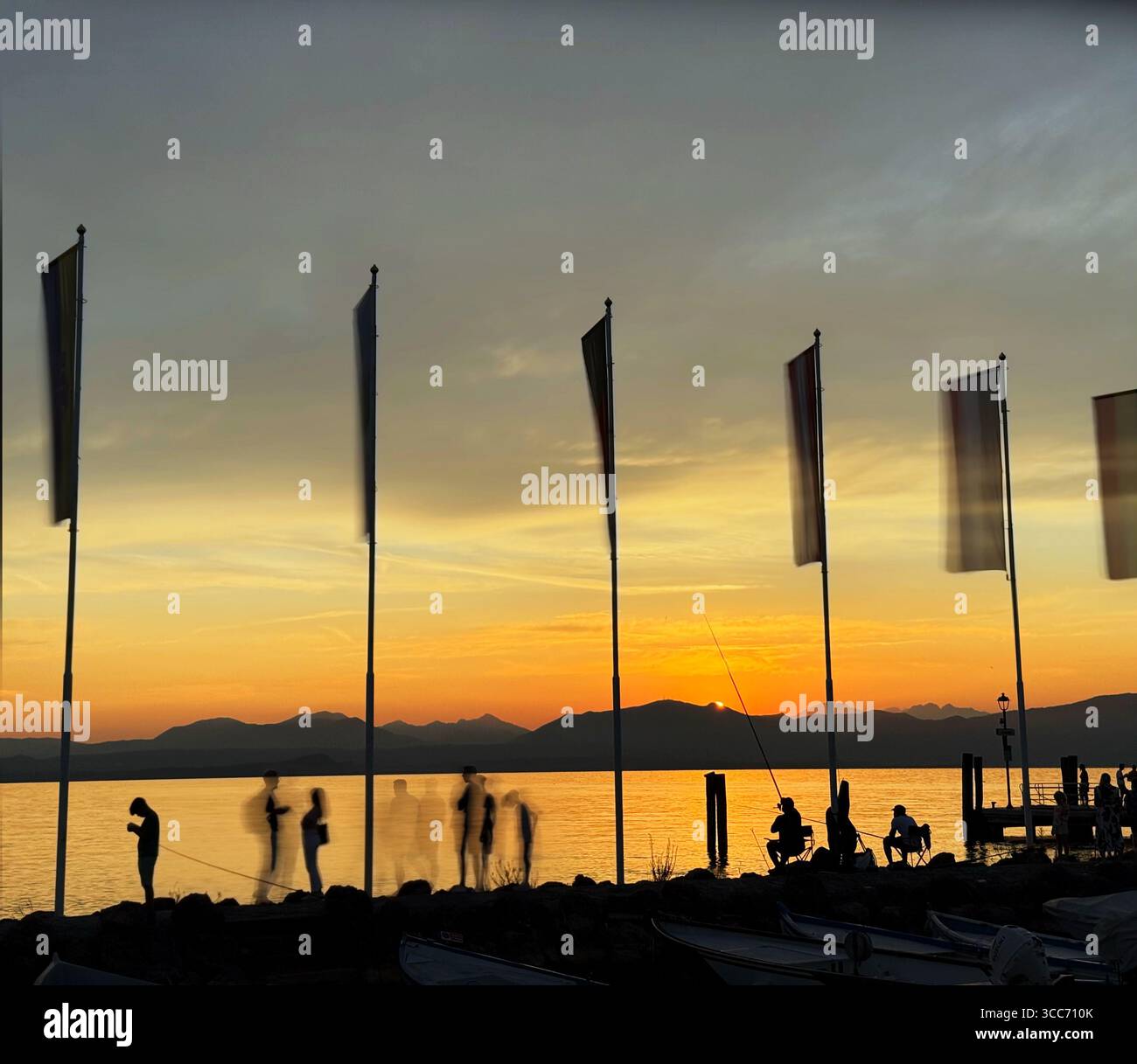 Cisano (near Bardolino) on Lake Garda, Italy. People gathered on harbour by flags for the sunset. - Smartphone Captured Stock Image
