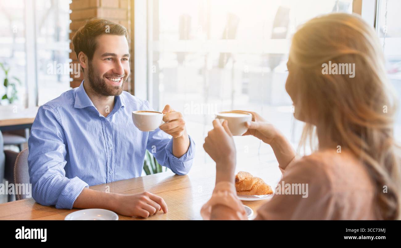 Cheerful young guy having coffee together with his girlfriend at cafe, chatting and smiling during breakfast Stock Photo