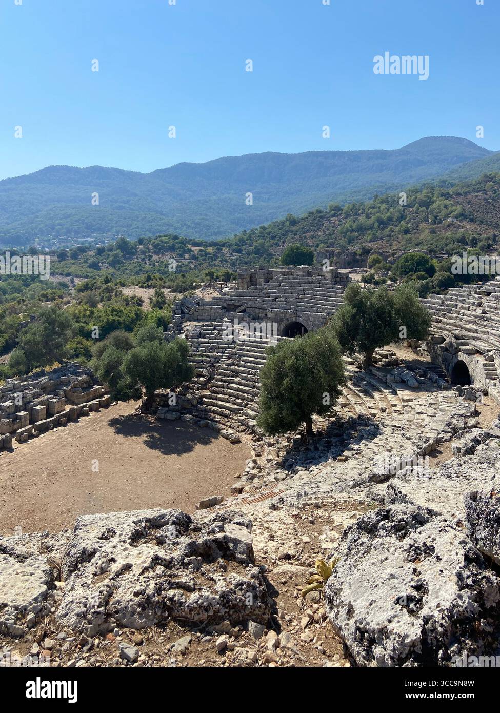 Ancient stone amphitheater nestled in a scenic mountainous landscape under a clear blue sky. - Smartphone Captured Stock Image