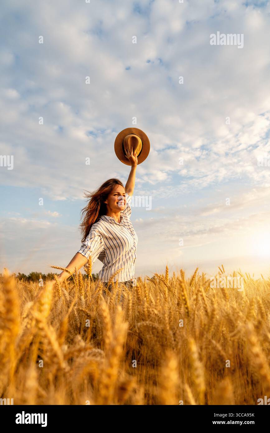 Free-spirited woman holding straw hat in raised hand standing in golden grain field, enjoying peaceful countryside Stock Photo