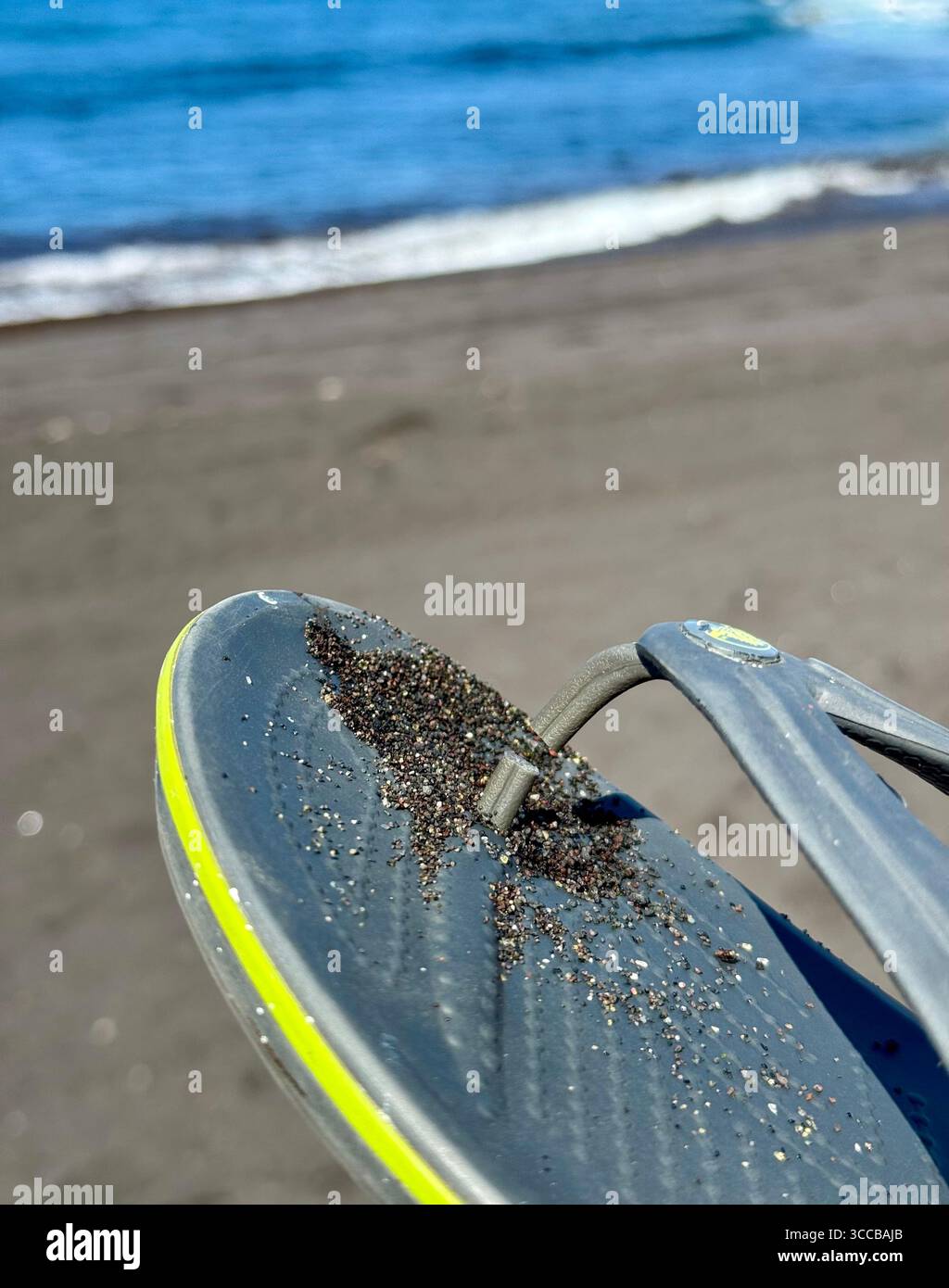 Close-up of torn flip-flops with black sand on a beach by the ocean. - Smartphone Captured Stock Image