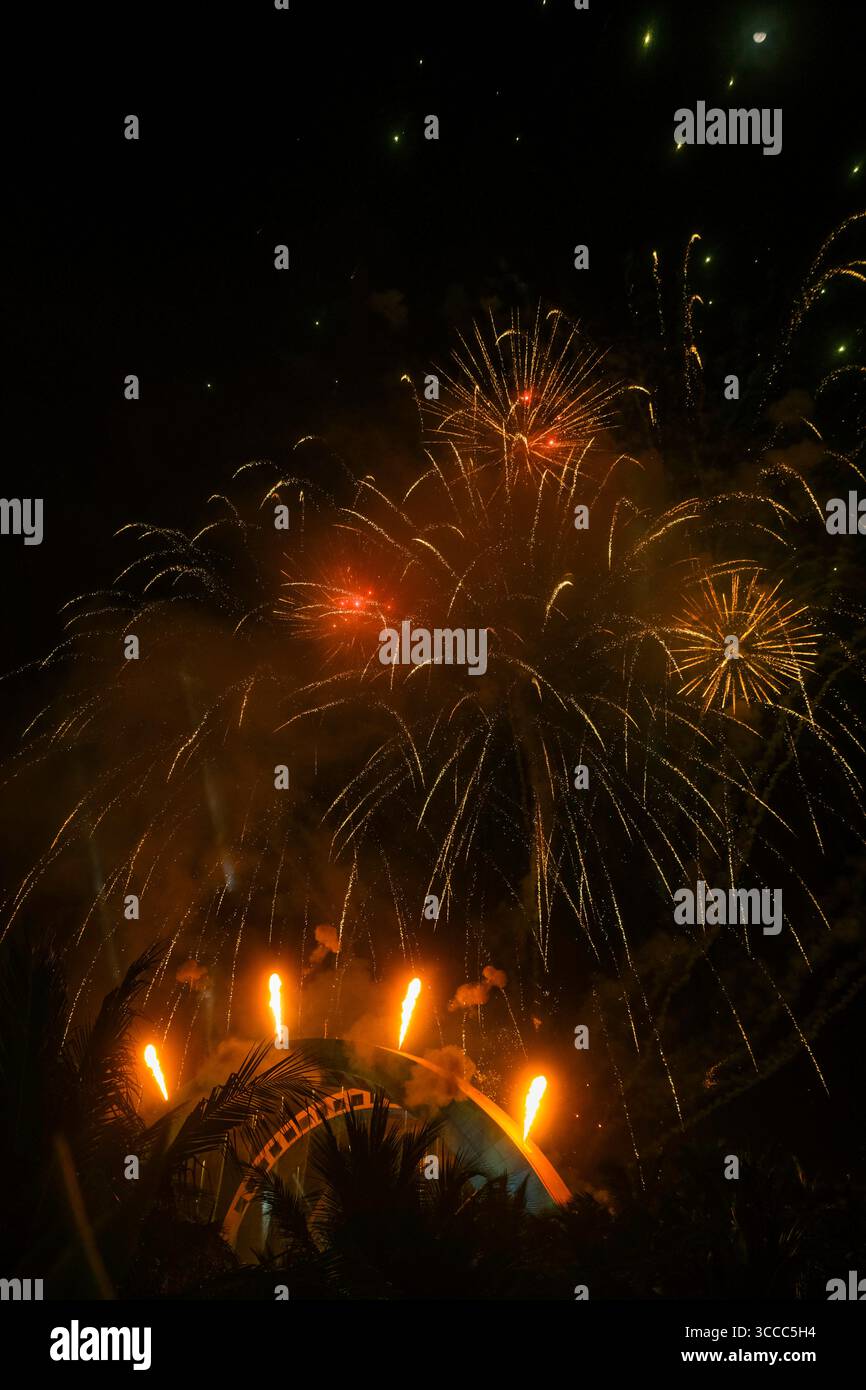 Golden fireworks burst above a dome stage with vertical fire jets and palm silhouettes, set against a dark night sky Stock Photo