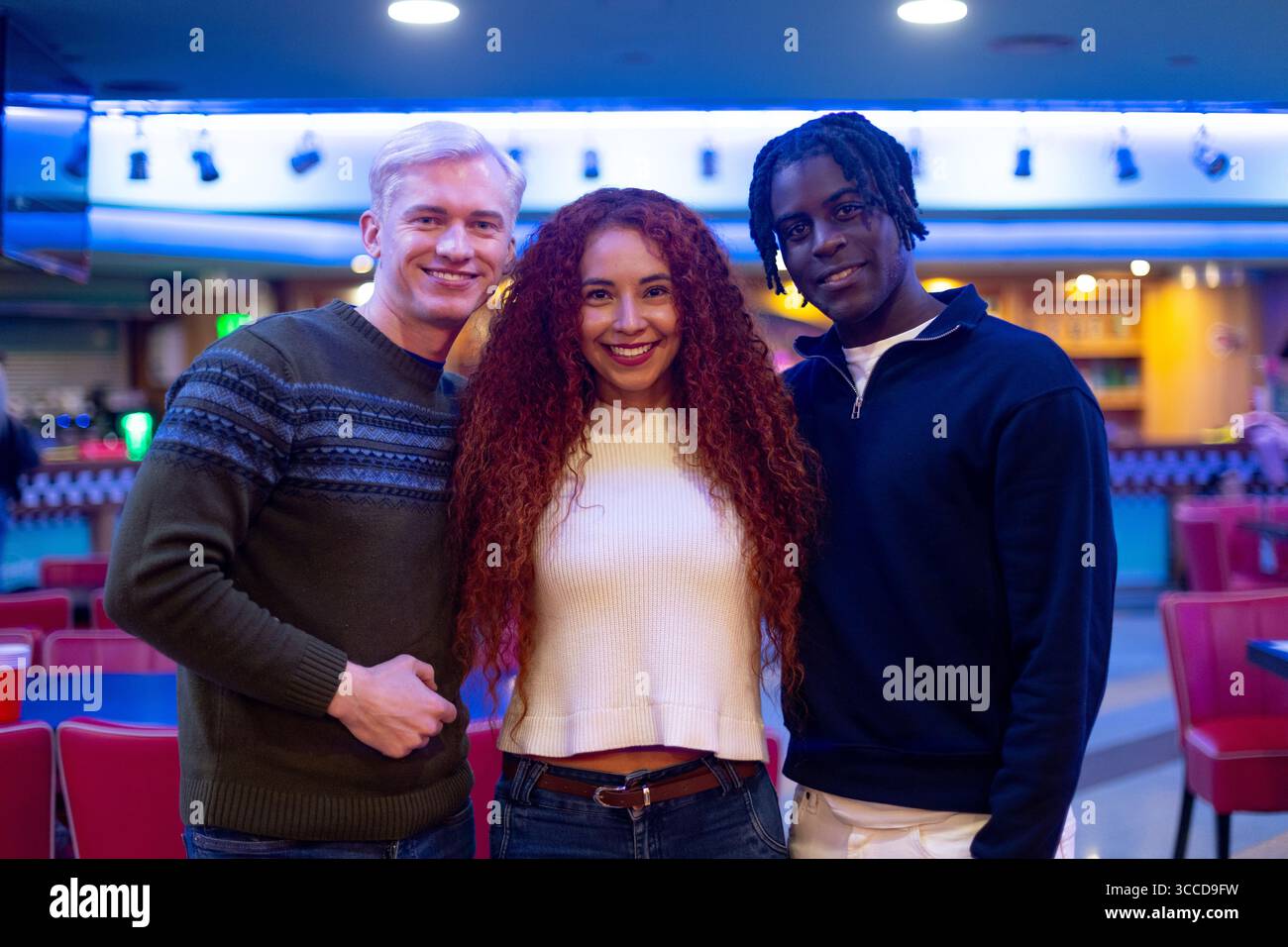 Three young adult friends smiling together inside a bowling alley, enjoying their free time together Stock Photo