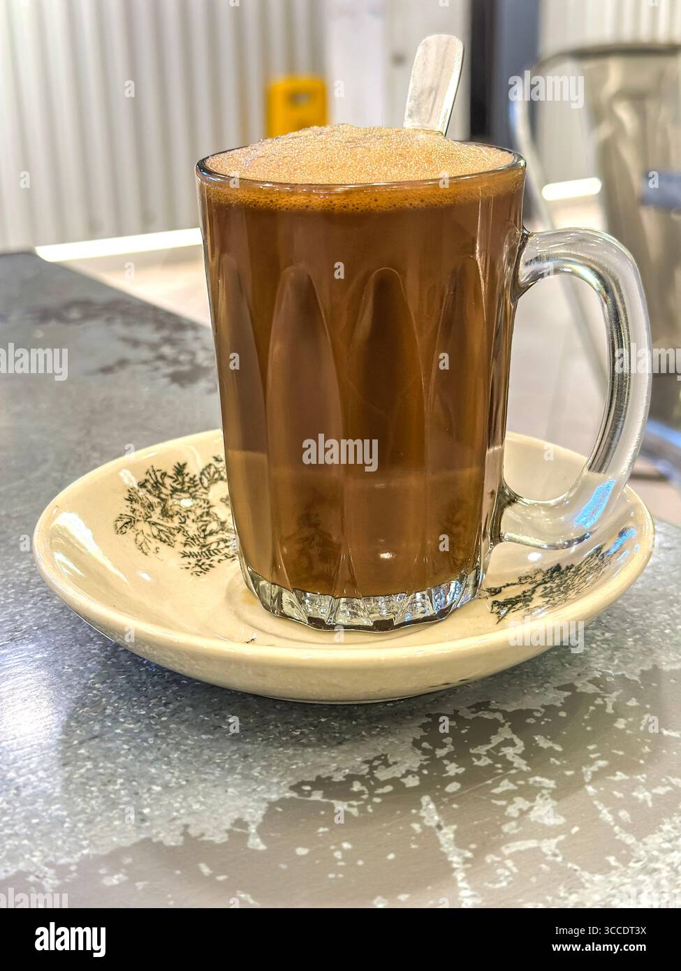 A frothy hot traditional hailam coffee in a transparent glass cup with spoon, on decorative saucer in a rustic kopitiam cafe kuala lumpur malaysia - Smartphone Captured Stock Image