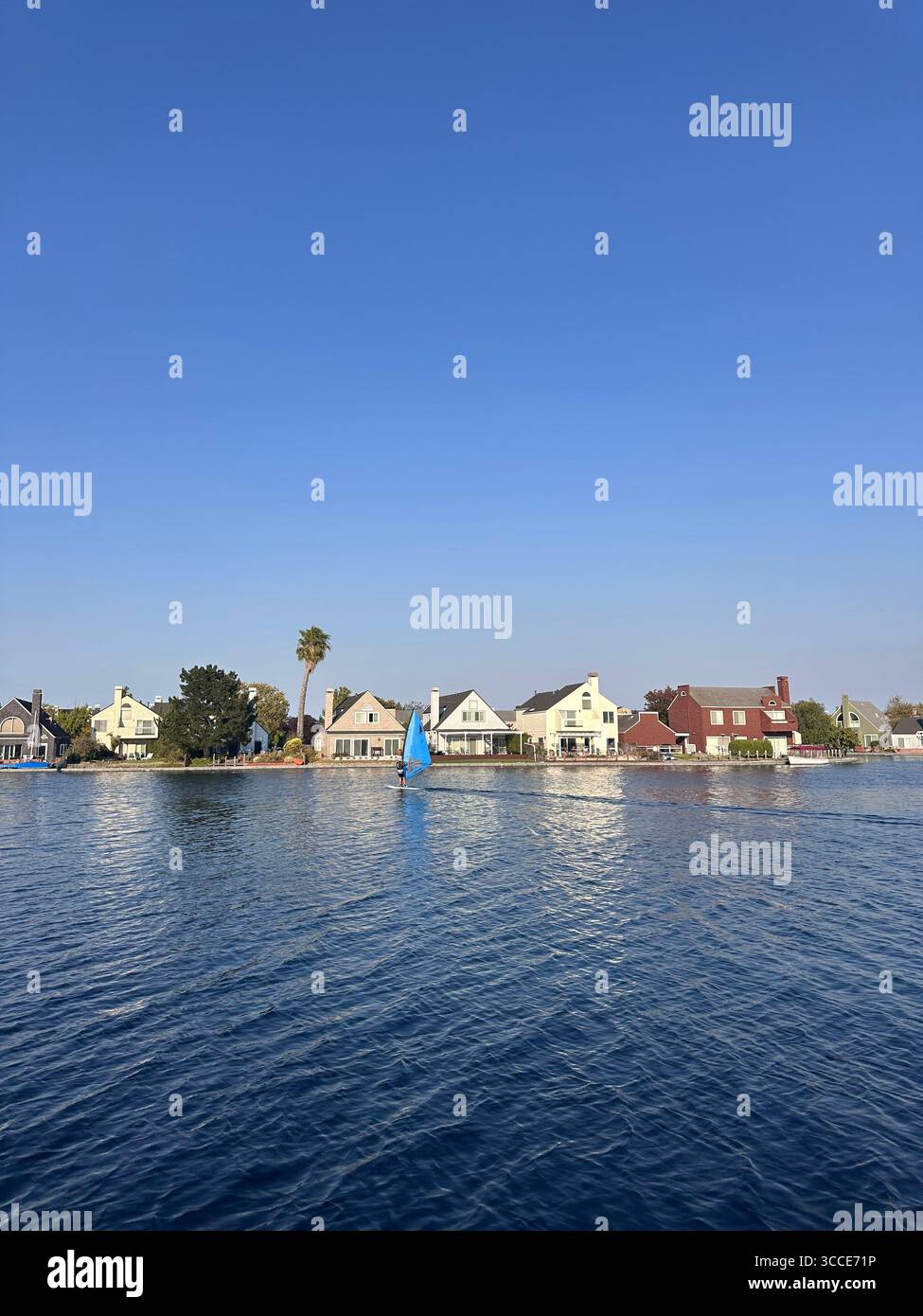 A windsurfer glides across a calm suburban lake in front of a row of colorful waterfront houses under a clear blue sky - Smartphone Captured Stock Image