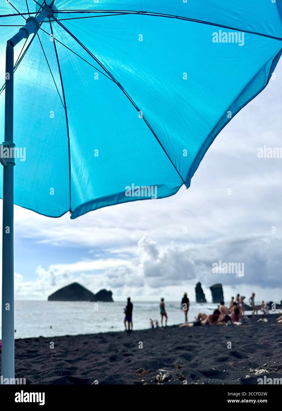 A blue beach umbrella providing shade at Mosteiros Beach in the Azores, with people enjoying the sun and the ocean view. - Smartphone Captured Stock Image