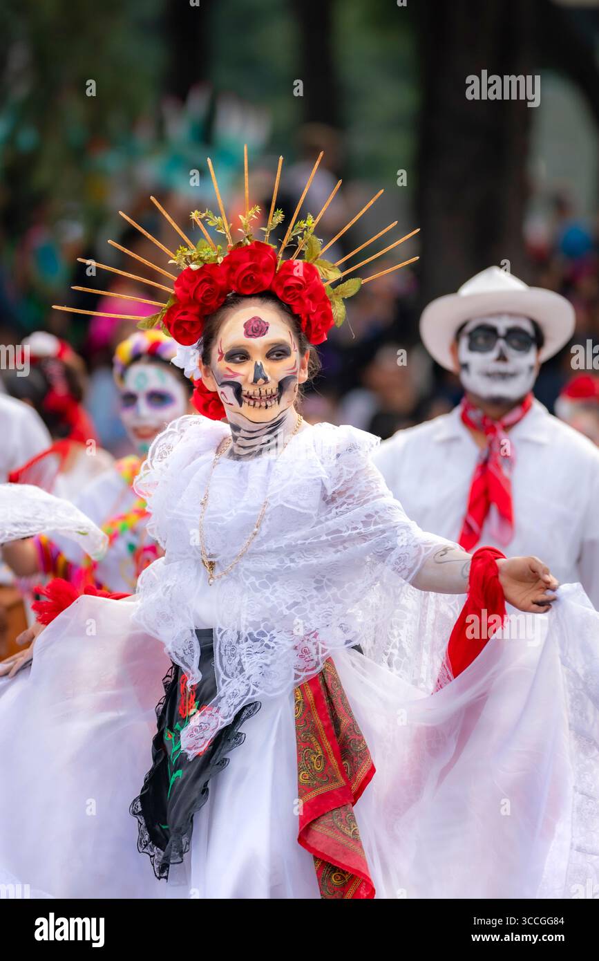 Mexico City, Mexico - November 2, 2024: Young Latina in white dress, skull makeup, and red floral crown parades through Mexico for the Day of the Dead Stock Photo
