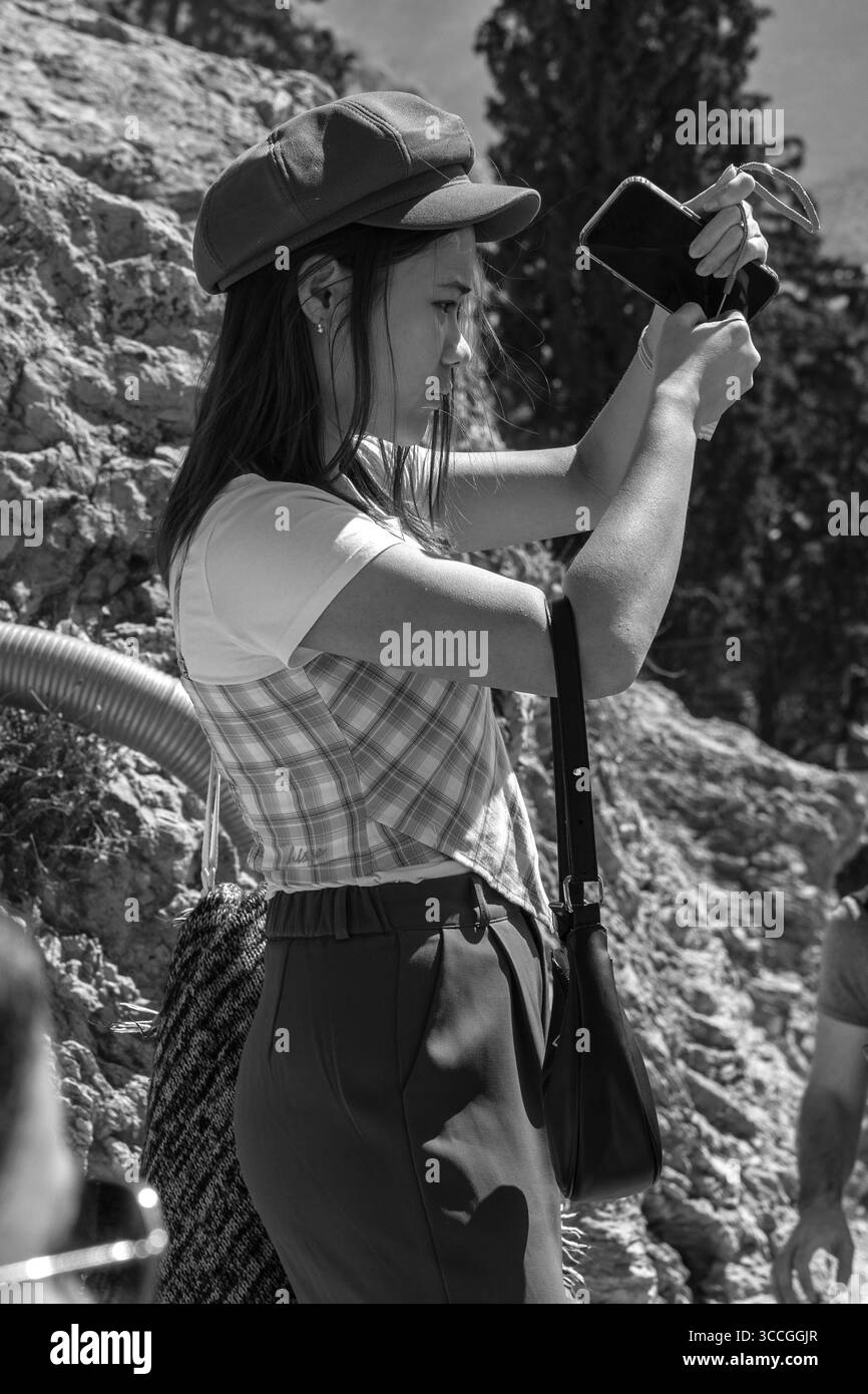 A woman stands at the Acropolis, Athens, using her phone to take photos on a hot summer day, surrounded by sunlit ancient ruins and city views. Stock Photo