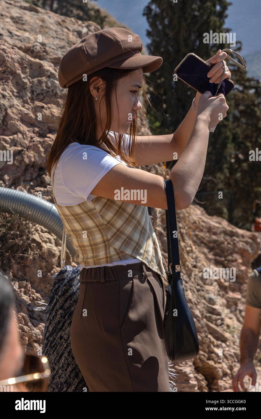 A woman stands at the Acropolis, Athens, using her phone to take photos on a hot summer day, surrounded by sunlit ancient ruins and city views. Stock Photo