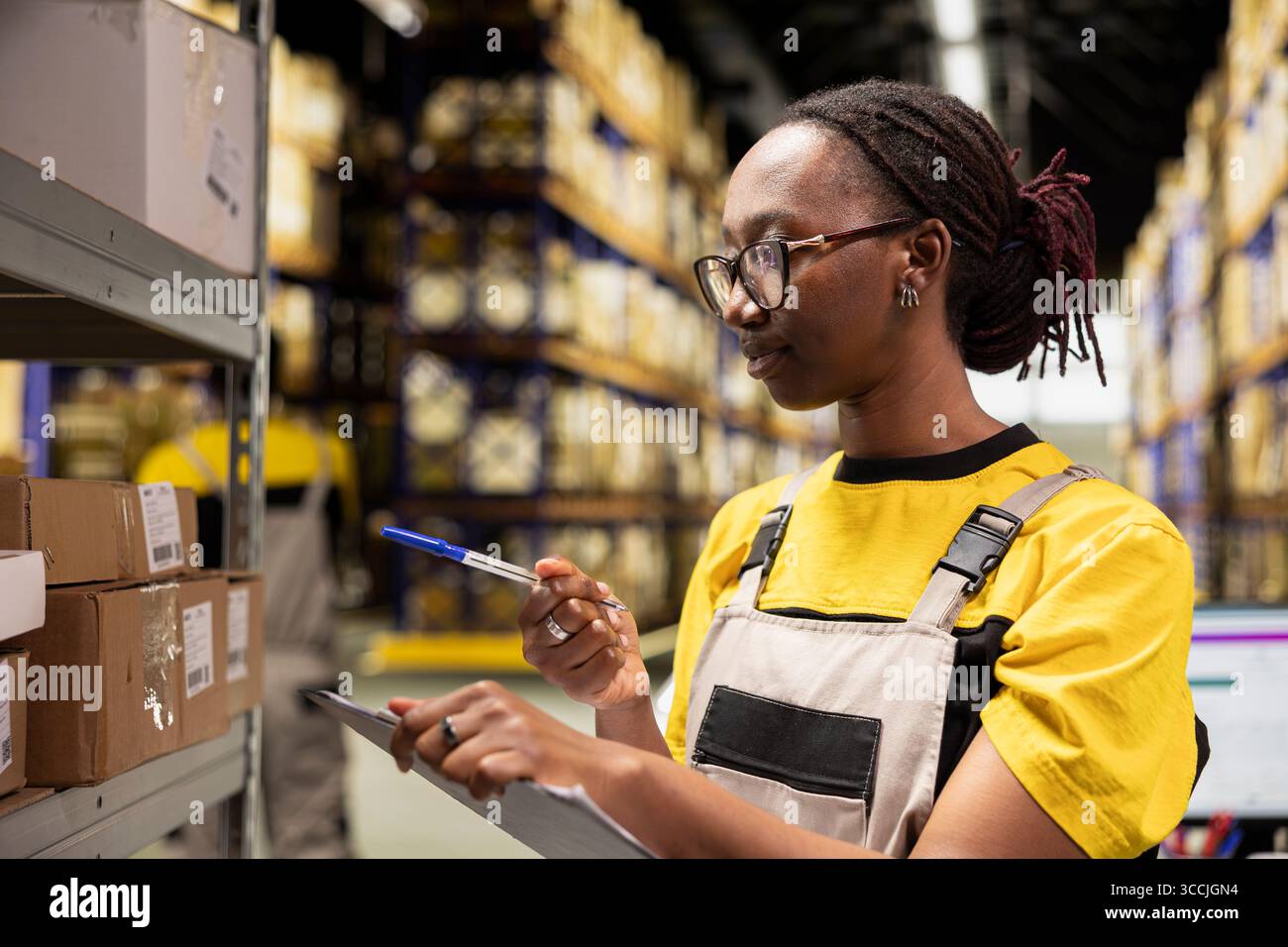 African american female worker checking tracking numbers tags placed on stacked pallet boxes in e-commerce warehouse. Woman handling order processing and parcels verification for shipment. Stock Photo