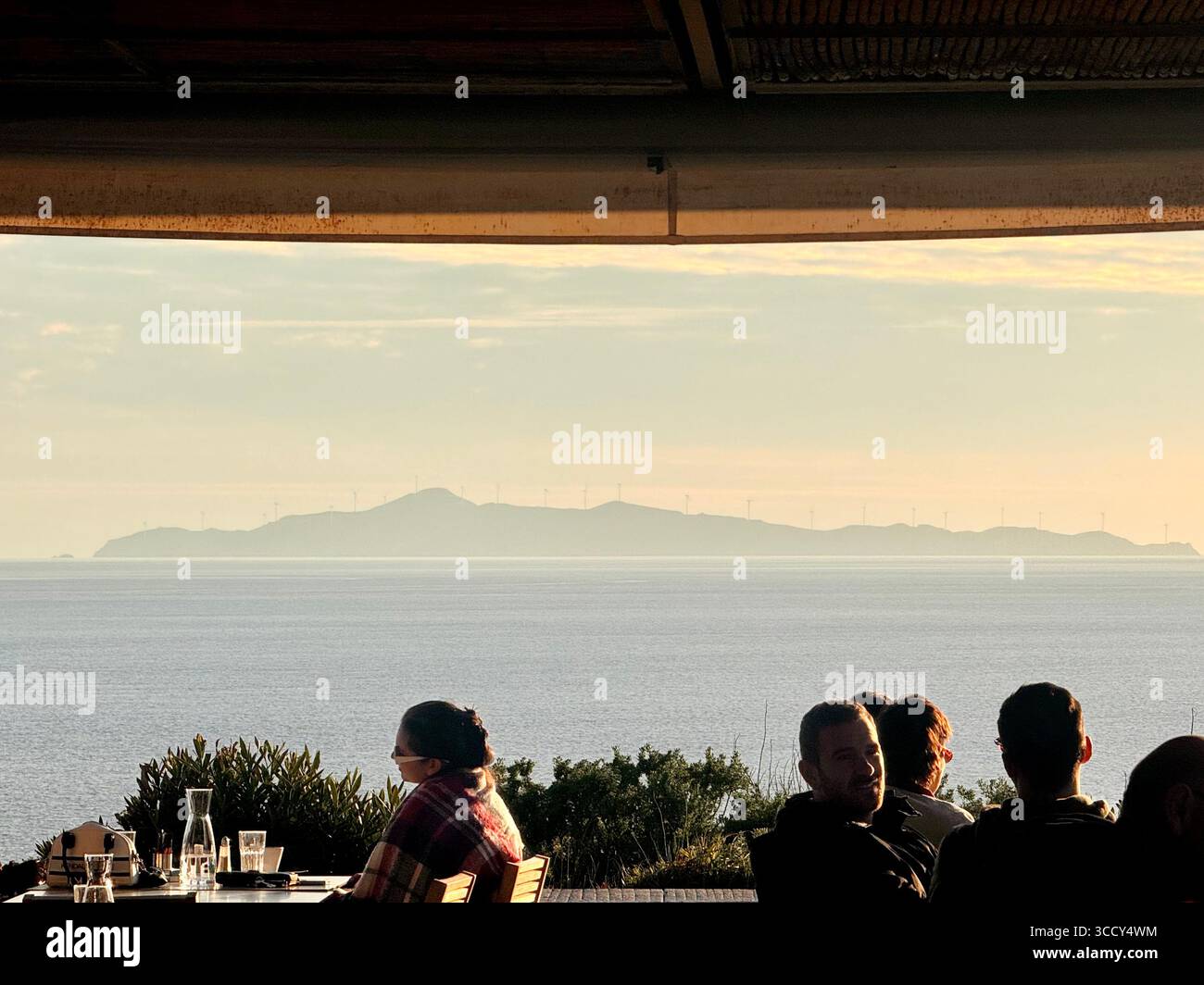 People dining on a scenic terrace with a view of Agios Georgios Island at sunset, Cape Sounion, Greece. - Smartphone Captured Stock Image