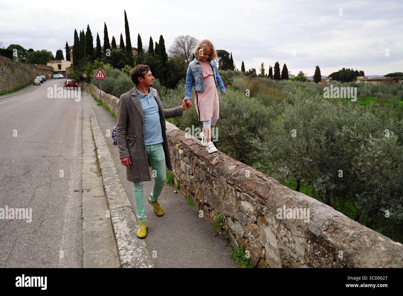 Father supports his daughter as she carefully walks along an ancient stone wall by the roadside. They enjoy a peaceful walk through the countryside, s Stock Photo