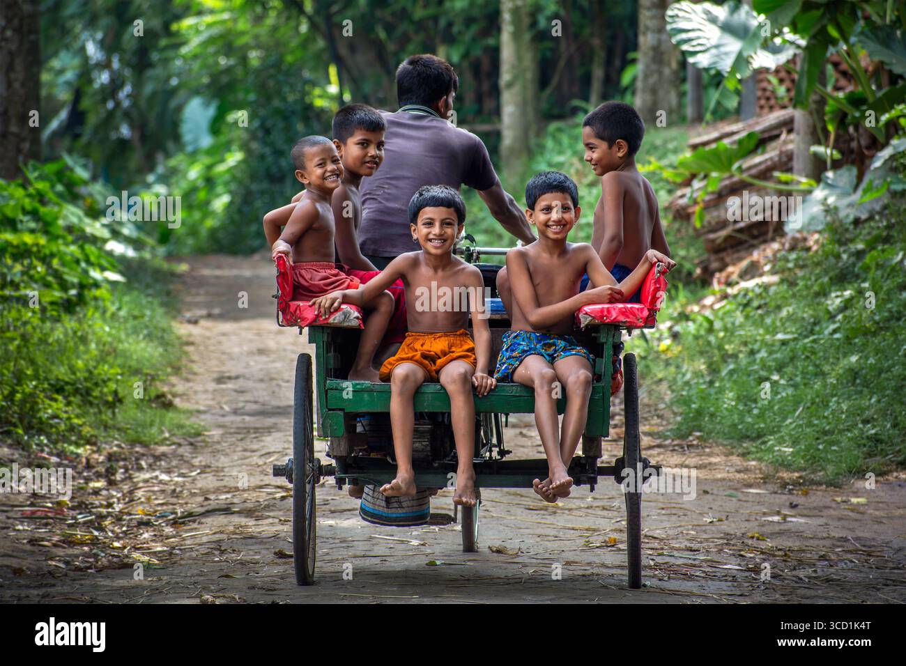 Bogura, Bangladesh - 23 August 2018: View of a vibrant green cart carrying smiling children, their joyous faces contrasting with the dusty path and lush foliage. Stock Photo