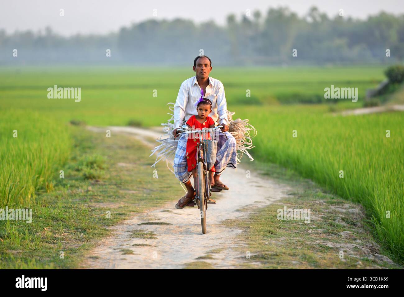 Bogura, Bangladesh - 04 April 2020: View of a father and child riding a bicycle along a dirt path flanked by vibrant green rice fields, creating a tranquil rural scene. Stock Photo