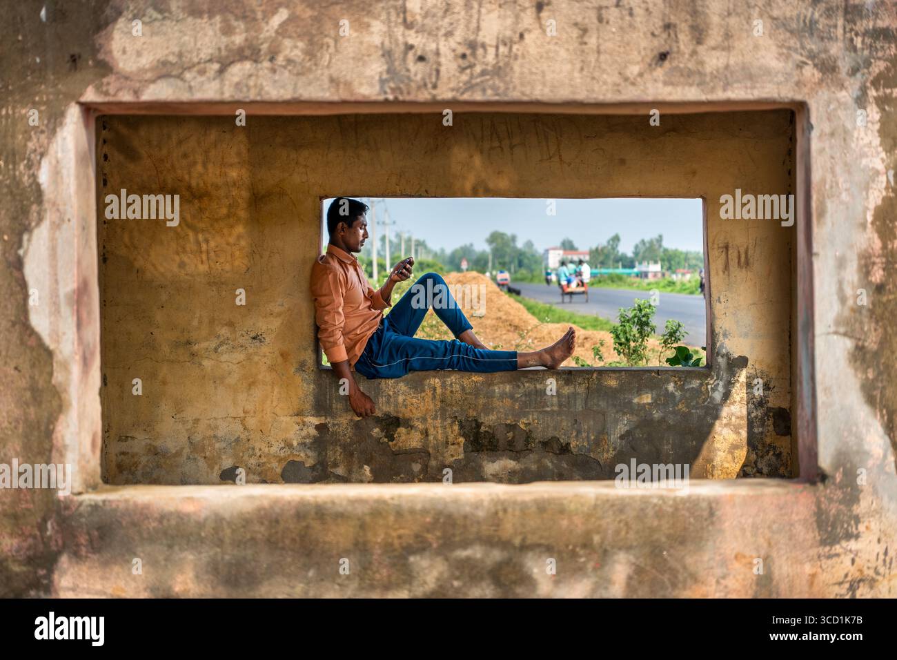 Bogura, Bangladesh - 04 June 2024: View of a young man sits contemplatively in a weathered window frame, gazing out at a highway, contrasting textures of old stone and vibrant greenery. Stock Photo