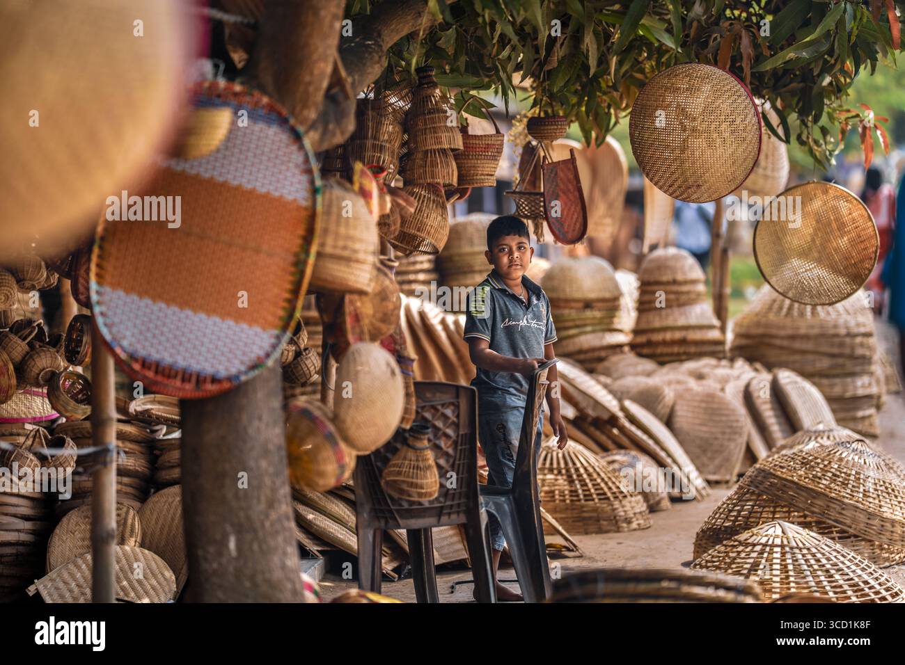 Bogura, Bangladesh - 27 February 2024: View of a local boy standing amidst a vibrant display of handcrafted bamboo and rattan products, showcasing the rich cultural heritage of the region. Stock Photo