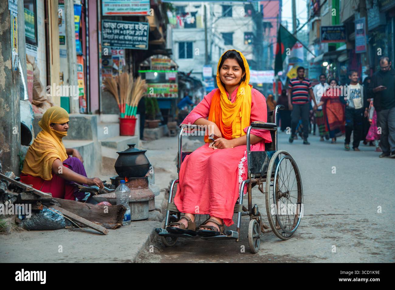 Bogura, Bangladesh - 17 December 2019: View of a smiling woman in a wheelchair amidst the bustling street scene, where vibrant colors meet the rustic charm of local life. Stock Photo