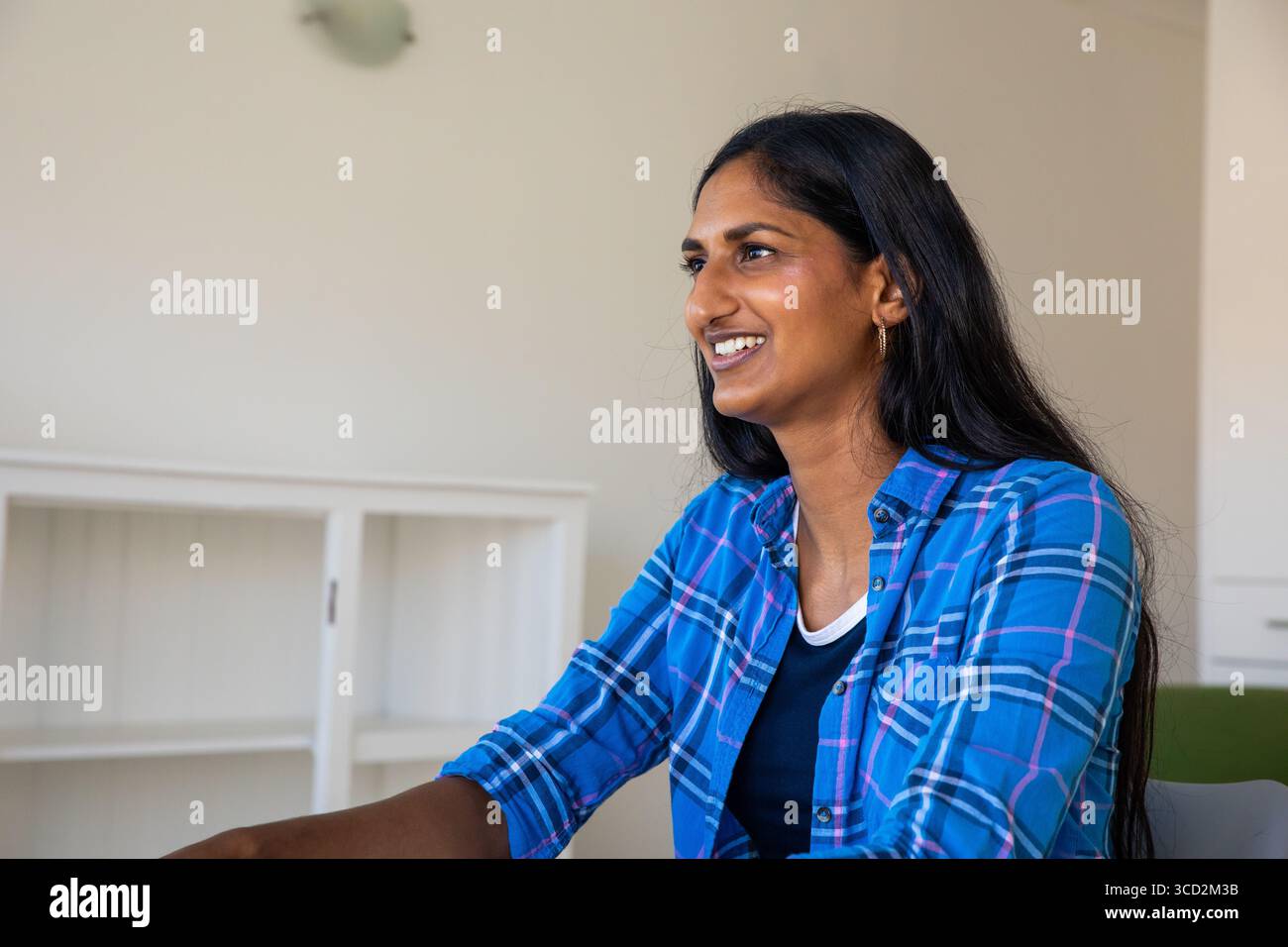 Indian woman sitting at desk wearing blue plaid shirt in office with ...