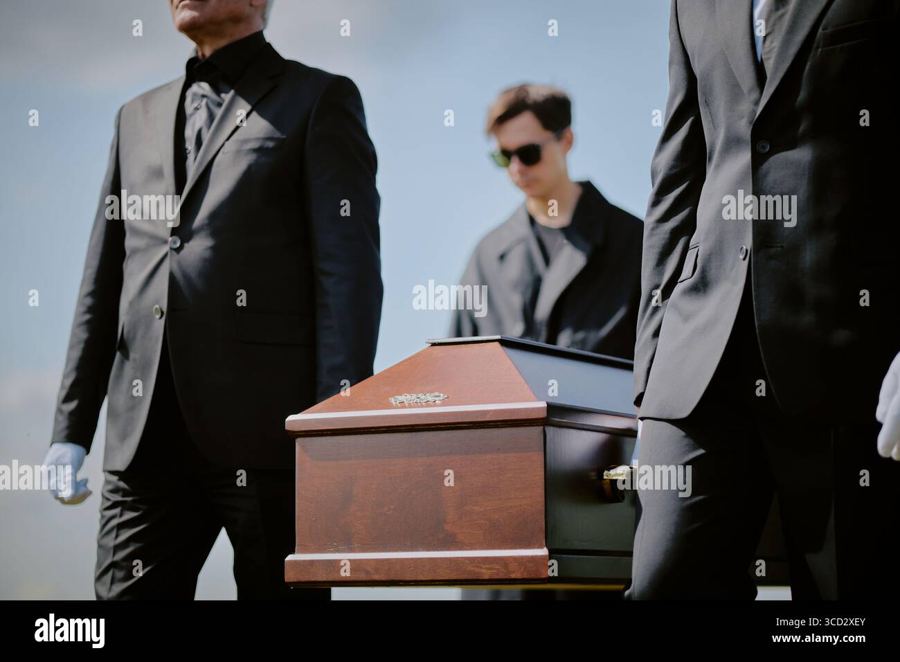 Caucasian middle aged men and young adult man wearing black suits carrying wooden coffin outdoors during funeral ceremony, young adult man wearing sunglasses walking behind casket Stock Photo