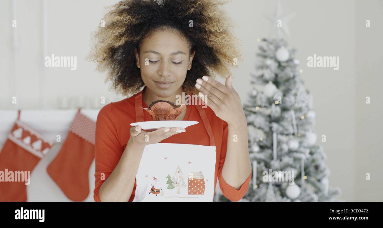 Young African cook with a freshly baked Christmas cake in her hand closing her eyes in bliss as she enjoys the aroma Christmas tree background Stock Photo