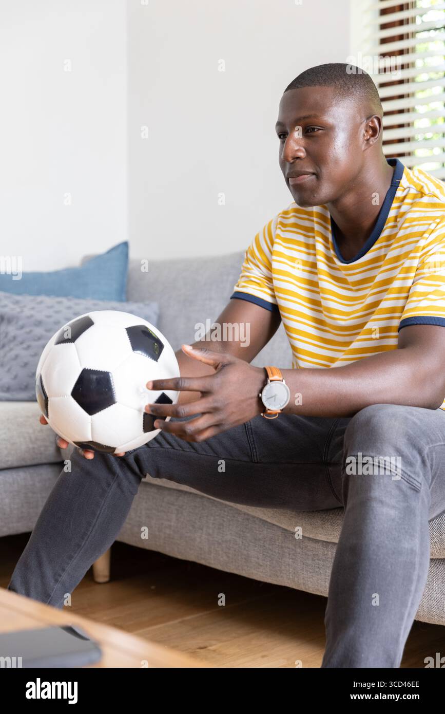 African American man sitting on sofa holding soccer ball in modern living room with tablet Stock Photo