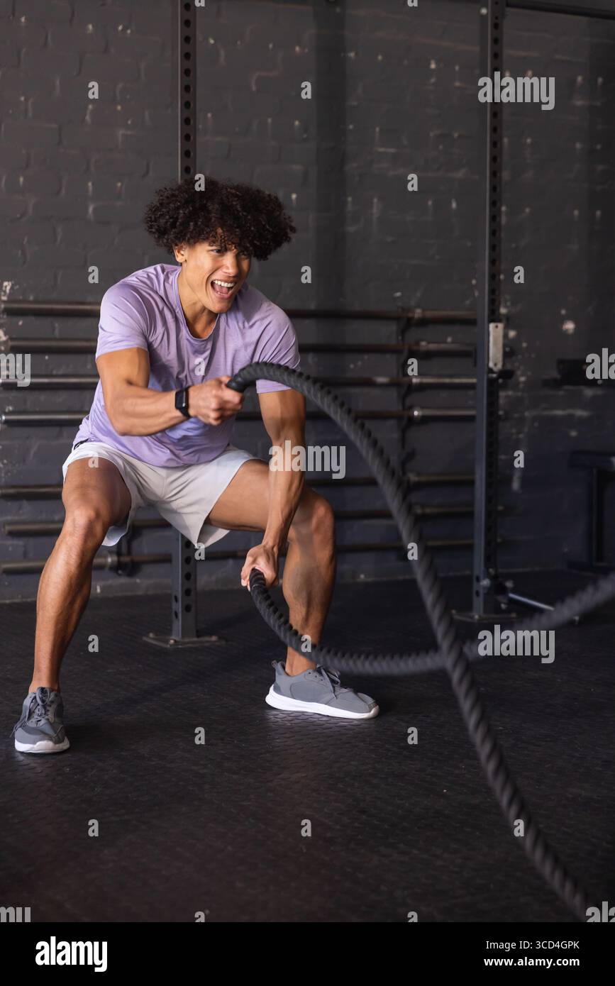 African American man wearing lavender shirt and gray shorts swinging battle ropes in gym studio Stock Photo