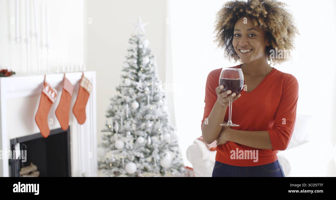 Attractive young African woman offering a Christmas toast as she raises a glass of red wine to the camera with a smile in front of a Christmas tree in Stock Photo