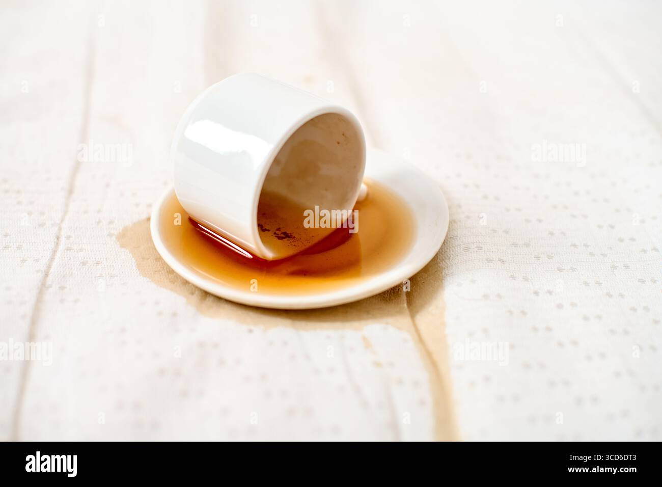 White cup overturned, coffee spilled on saucer and fabric. Soft natural lighting highlights liquid trail, creating casual yet dramatic contrast. Simpl Stock Photo