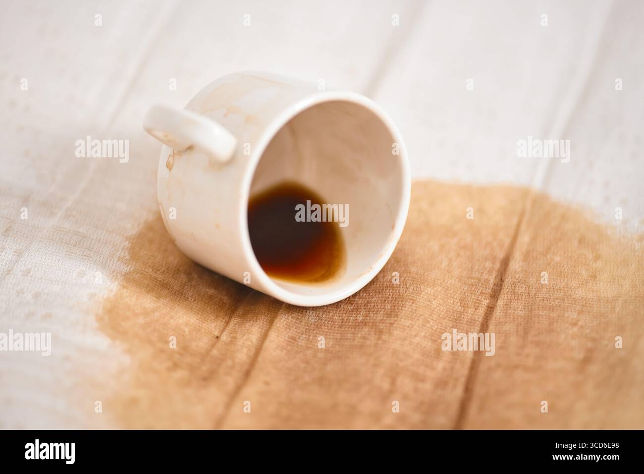 White ceramic cup resting on side with small coffee puddle on light fabric. Simple composition emphasizes spill, creating contrast between dark coffee Stock Photo