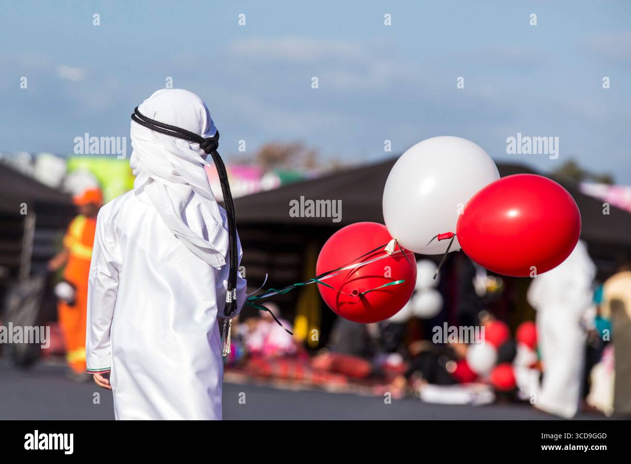Arab child wearing traditional white attire holding red and white balloons during outdoor festival celebration, Middle Eastern culture and heritage. Stock Photo