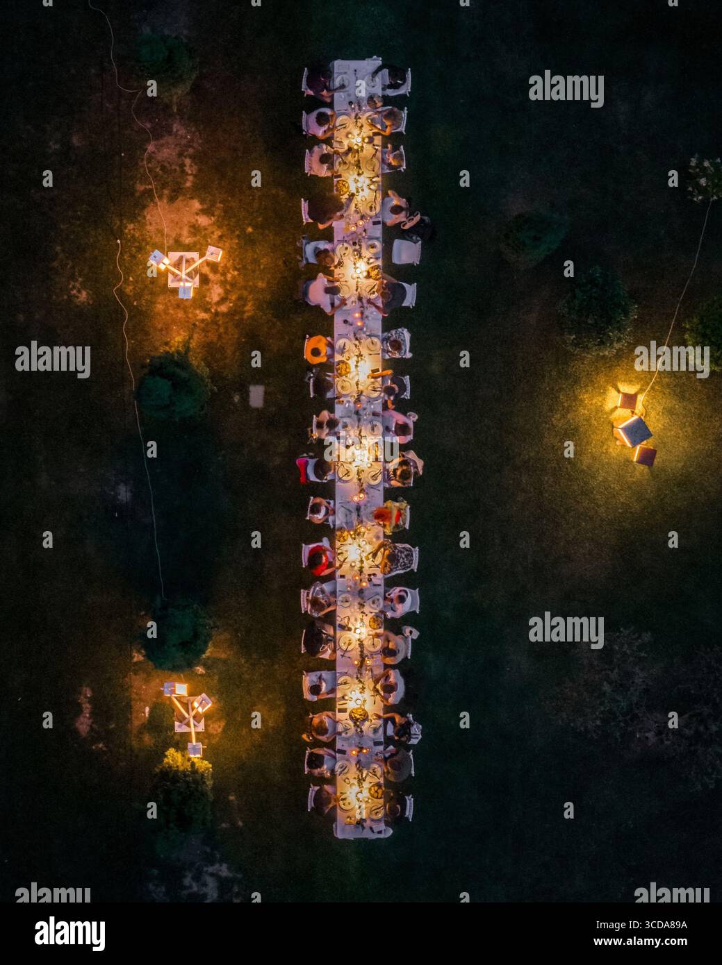 Aerial view of a long table set for a warm, inviting dinner under the soft glow of lights in the dark, green lawn, Assisi, Umbria, Italy. Stock Photo
