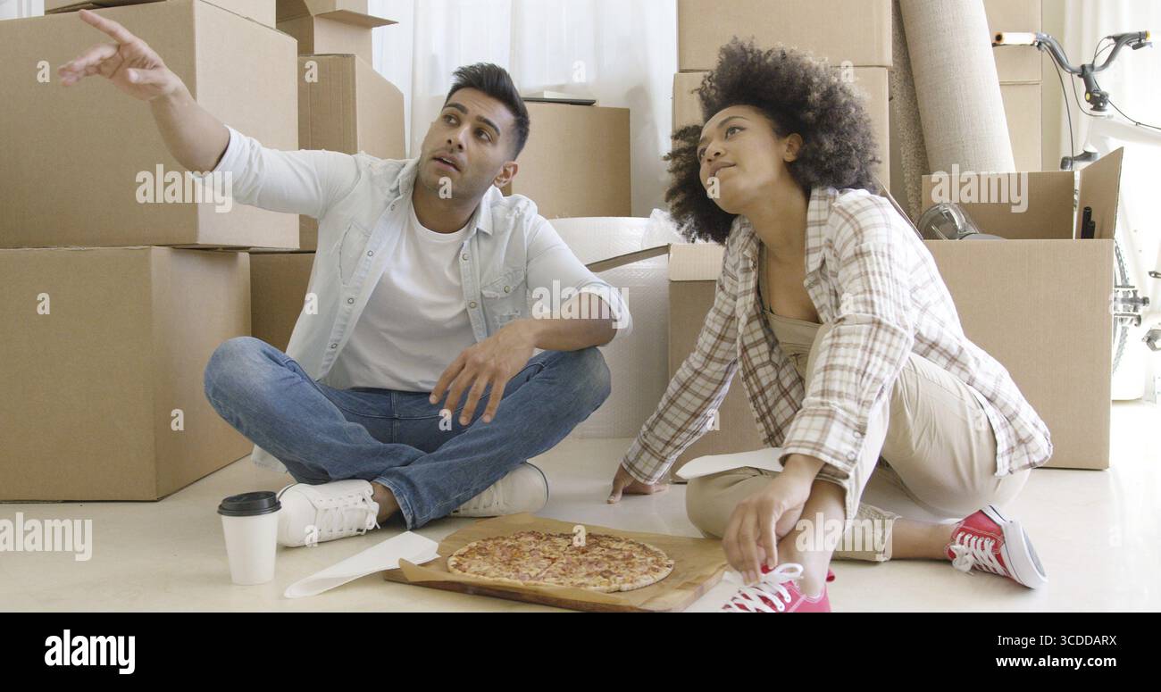Young couple eating pizza on the floor in front of a stack of brown packing boxes as they move house and chatting pointing to something off screen Stock Photo