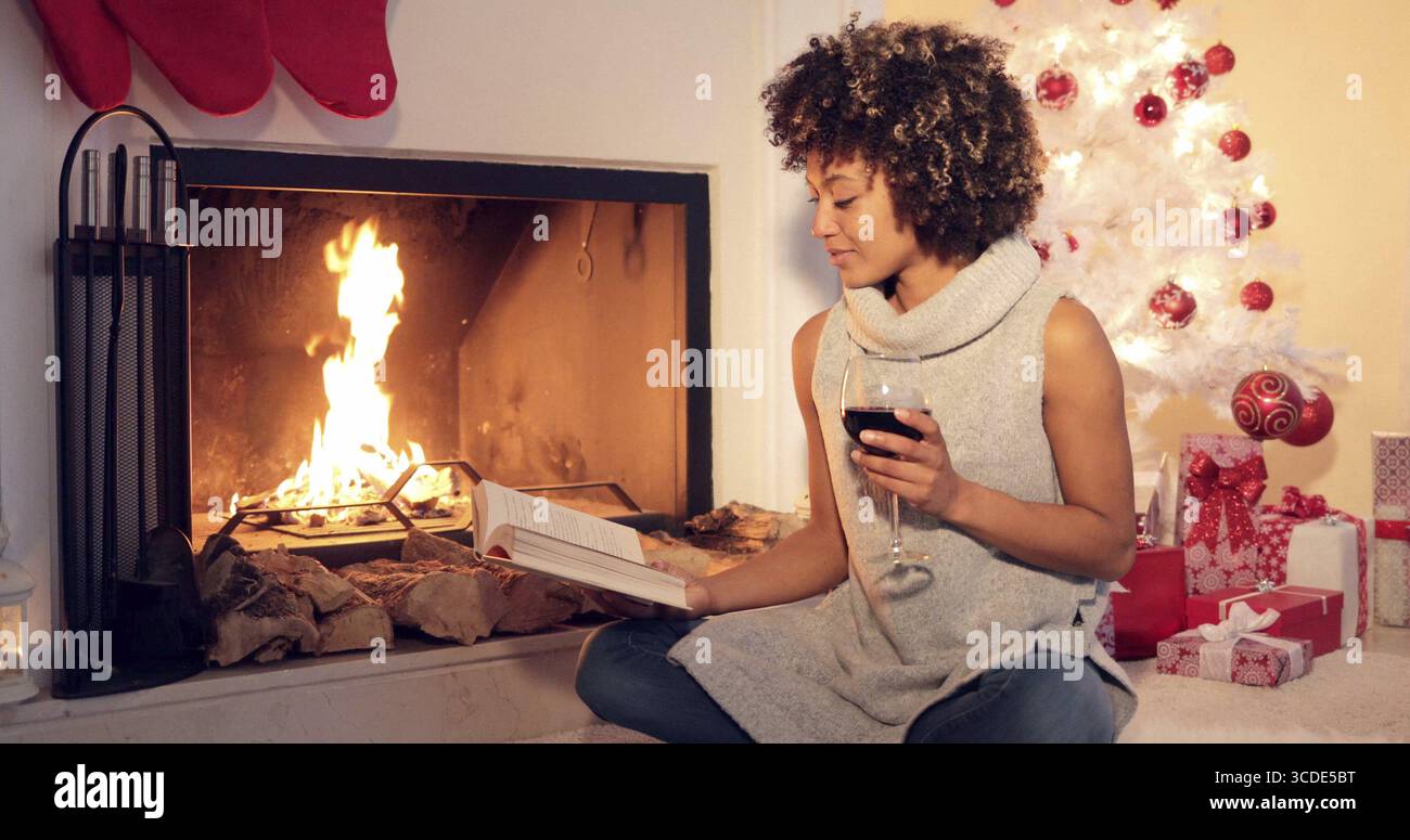 Young woman reading a drinking red wine as she relaxes in front of a blazing fire alongside a Christmas tree and gifts Stock Photo