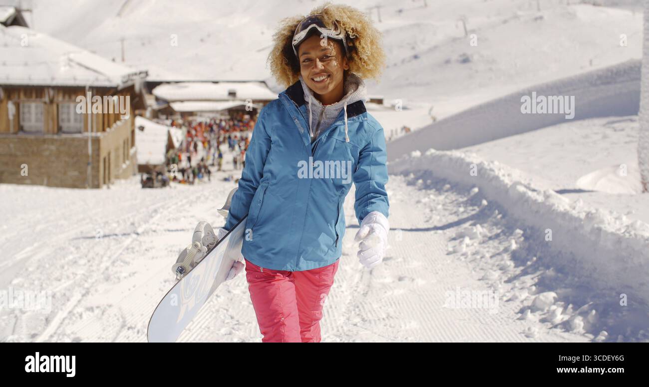 Smiling pretty young woman with a trendy afro hairstyle carrying a snowboard standing in fresh winter snow at a mountain ski resort Stock Photo
