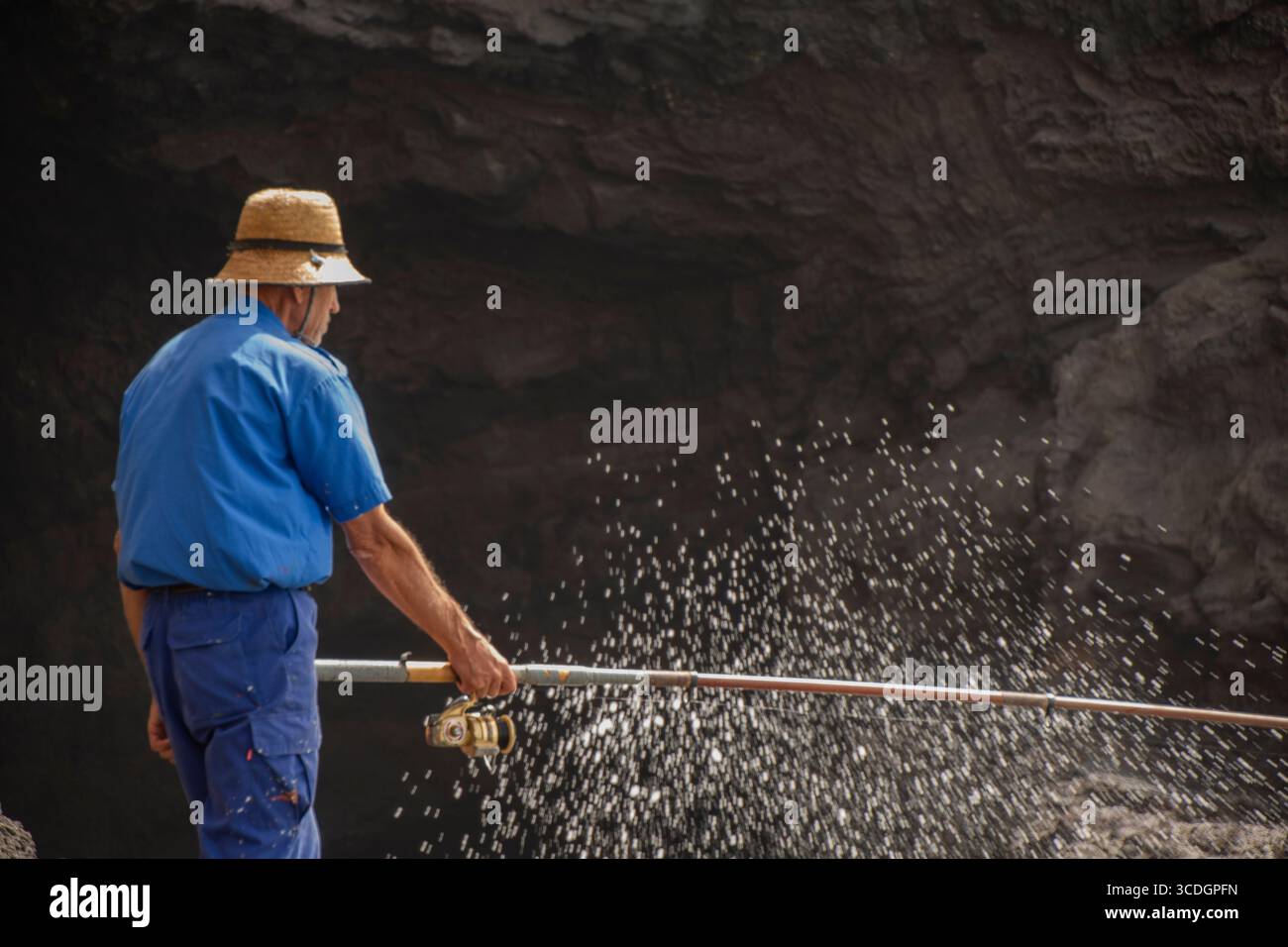 Hombre pescando con caña en una rocosa costa oceánica, olas rompiendo, vestido con traje azul y sombrero de paja, captura la esencia de la pesca Stock Photo
