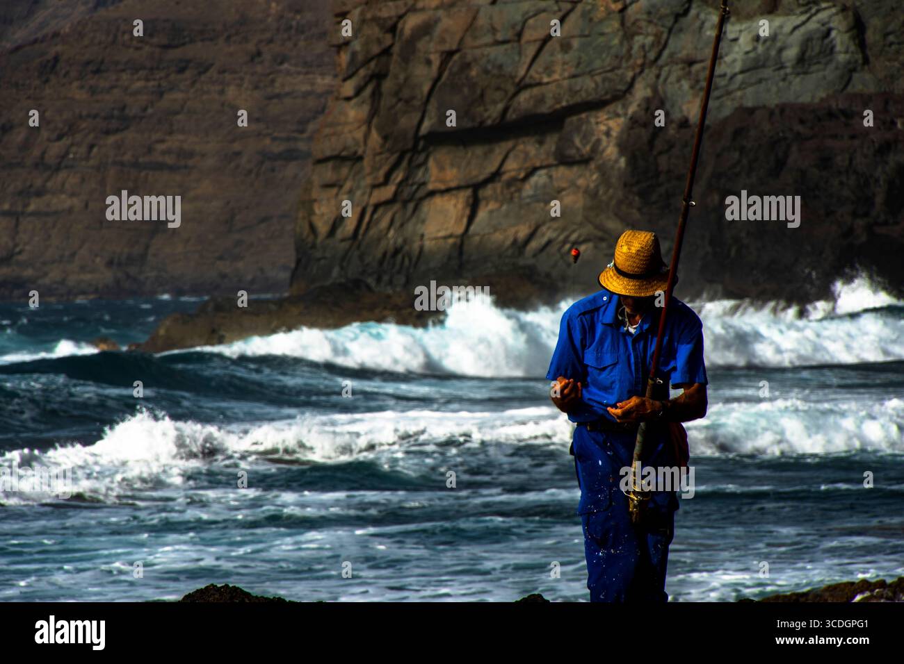 Pescador tradicional con caña junto a la costa rocosa del océano Atlántico, vistiendo ropa azul y sombrero de paja, frente a un paisaje marino con ola Stock Photo