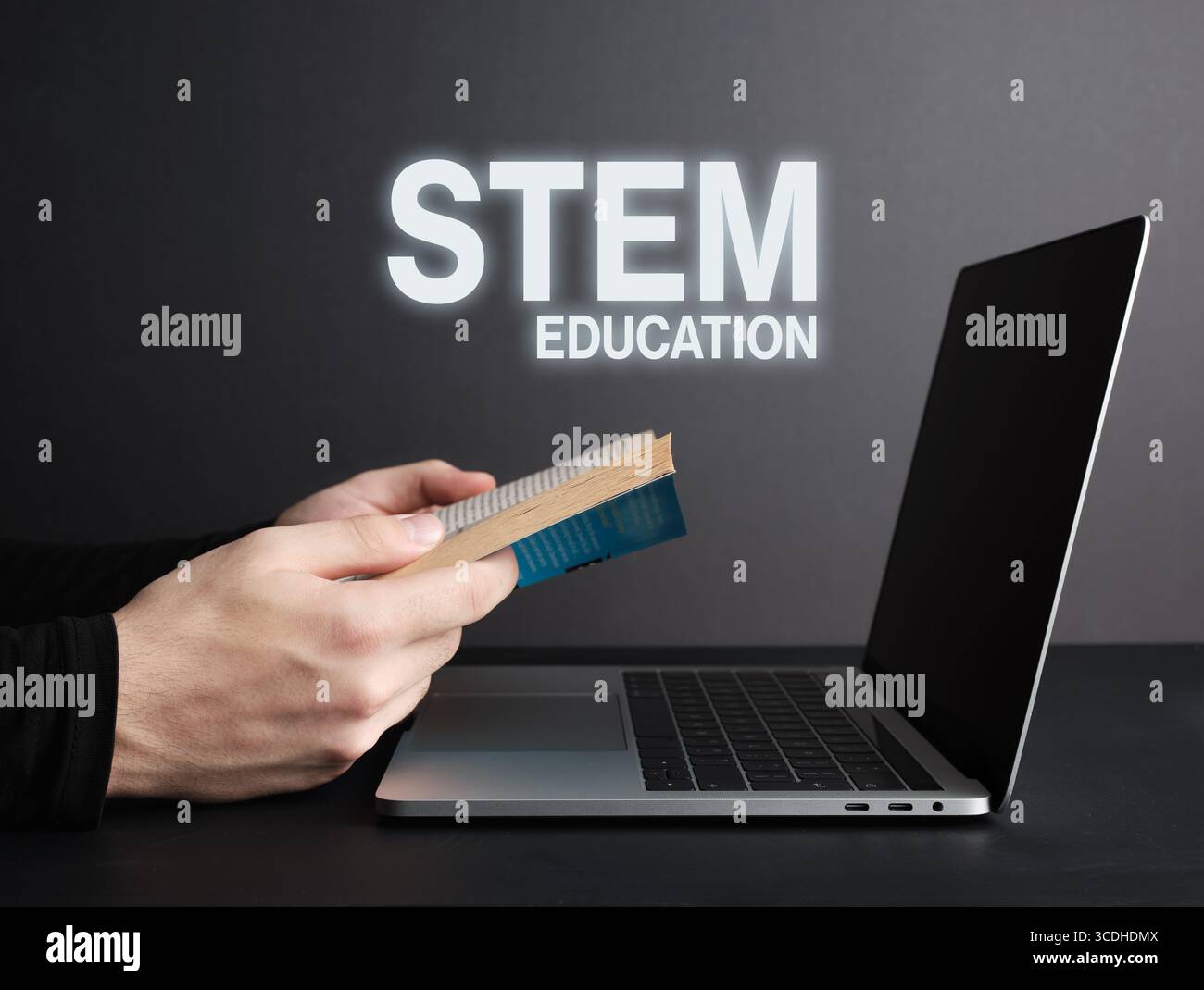 Hands holding an open book beside a laptop, featuring STEM education text against a dark gray background, emphasizing learning and innovation Stock Photo