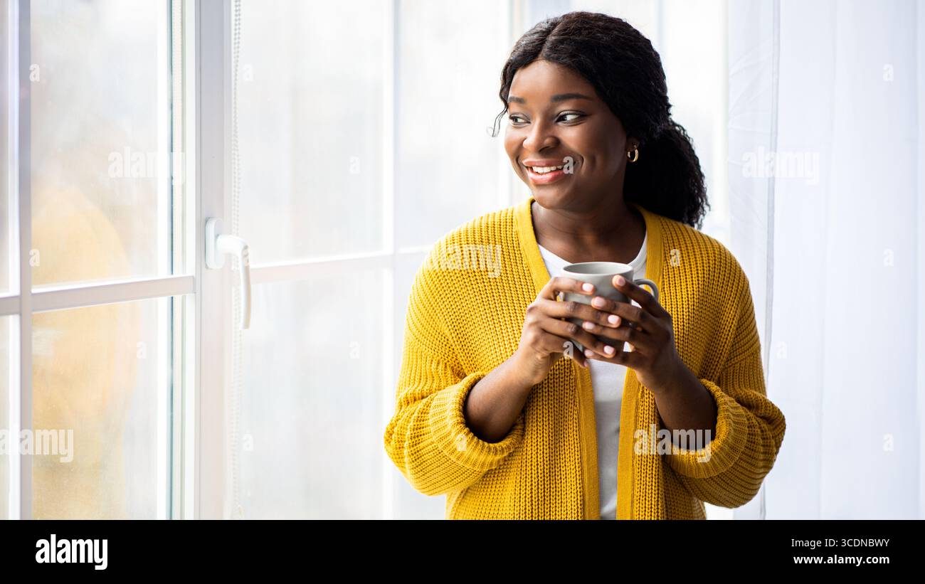 Black lady standing looking through window, drinking tea Stock Photo